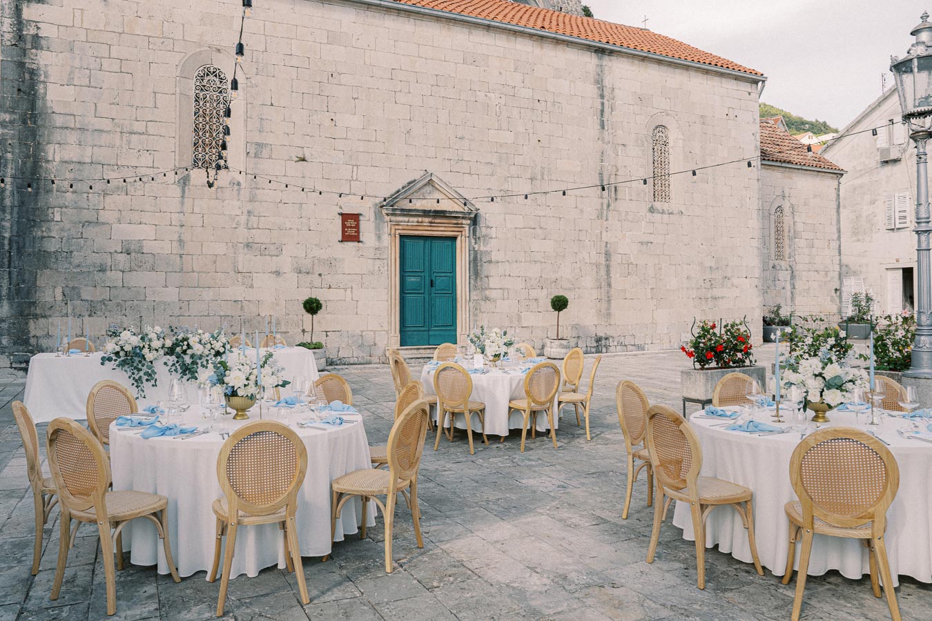 Outdoor wedding reception setup with round tables draped in white tablecloths, elegant floral centerpieces, and wooden chairs, set against a historic stone building with string lights.