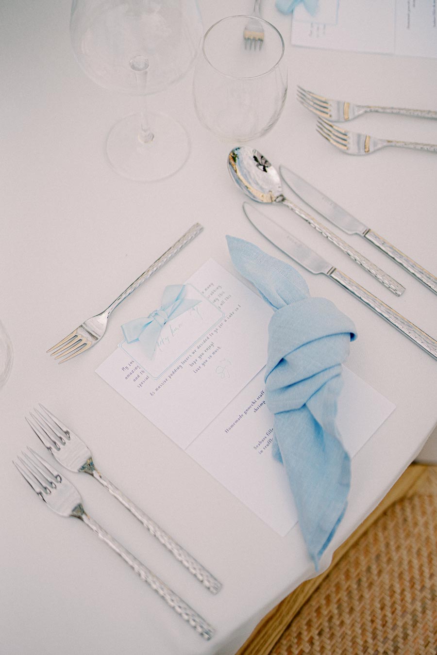 Elegant wedding table setting with silver cutlery, blue folded napkin, and menu card on a white tablecloth.