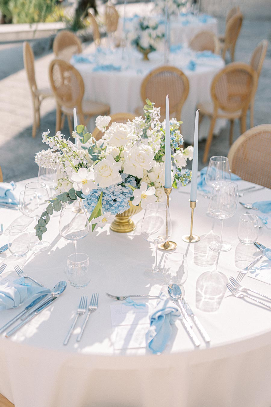 Elegant outdoor wedding reception table setting with white tablecloth, featuring a floral centerpiece of white roses and blue flowers, complemented by tall candlesticks and neatly arranged cutlery.