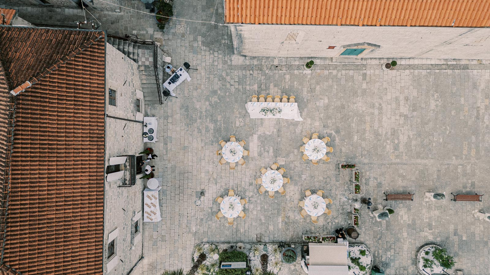 Aerial view of a beautifully arranged outdoor wedding venue with round tables set for guests, located in a stone-paved courtyard surrounded by historic buildings with orange-tiled roofs.