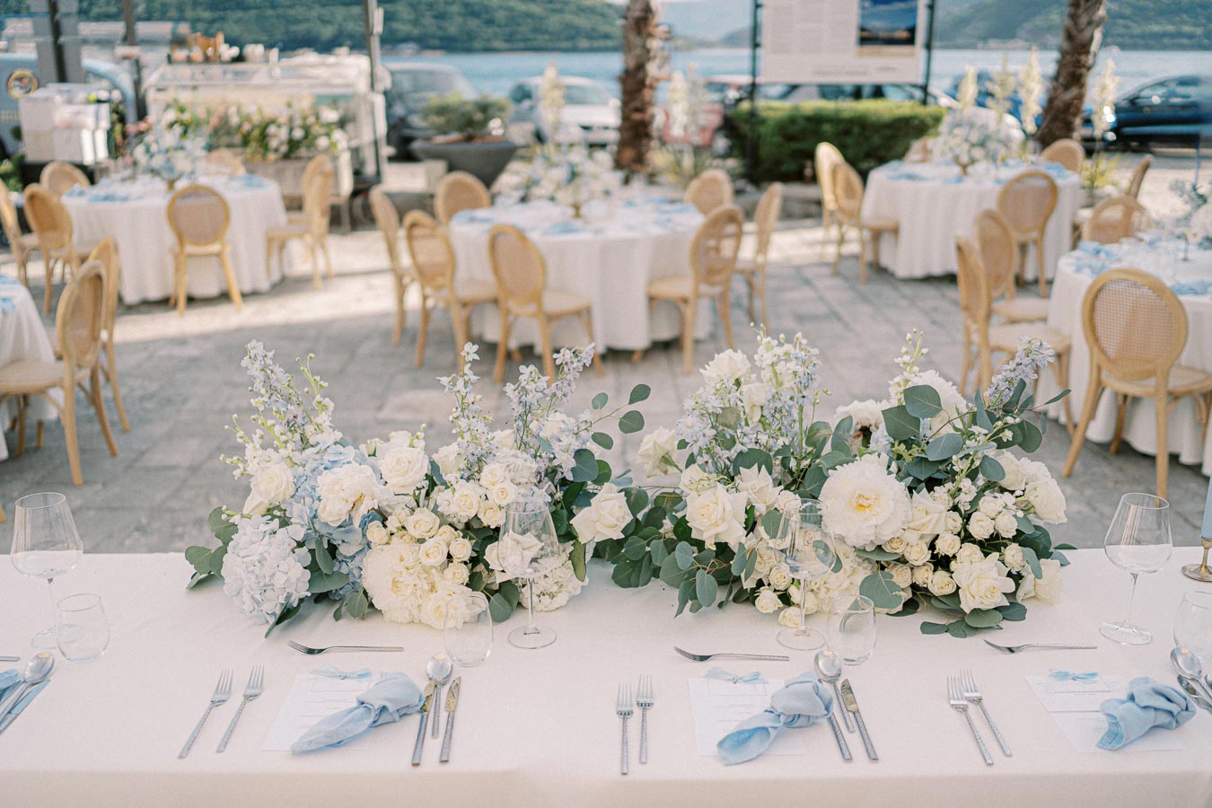 Elegant outdoor wedding reception setup with beautifully arranged floral centerpieces, featuring white and pastel flowers, surrounded by round tables and wooden chairs, overlooking a scenic waterfront backdrop.
