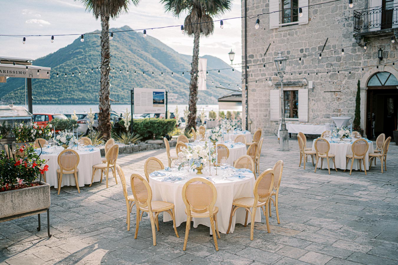 Outdoor wedding reception setup with elegantly decorated tables and chairs on a stone patio, surrounded by string lights and overlooking a scenic mountain and lake view.