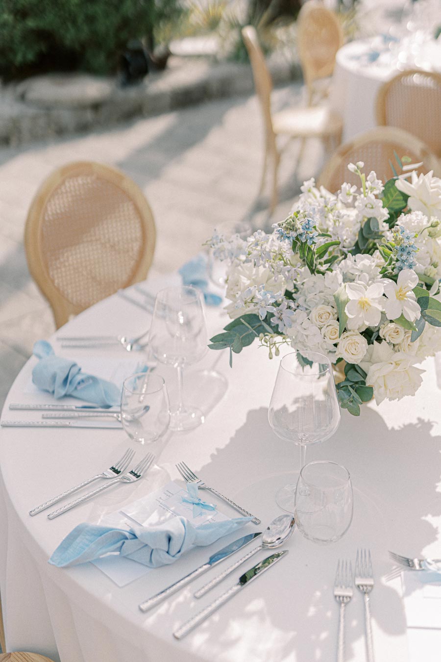 Elegant outdoor wedding table setting with a white tablecloth, blue napkins, and a centerpiece of white and blue flowers, surrounded by wicker chairs.