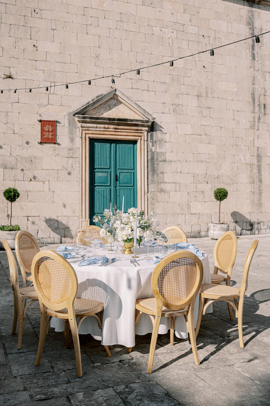 Elegant outdoor dining setup with a round table covered in a white tablecloth, surrounded by wooden chairs. The table is adorned with a floral centerpiece and set against a historic stone building with a teal door and string lights above, creating a charming and romantic atmosphere.