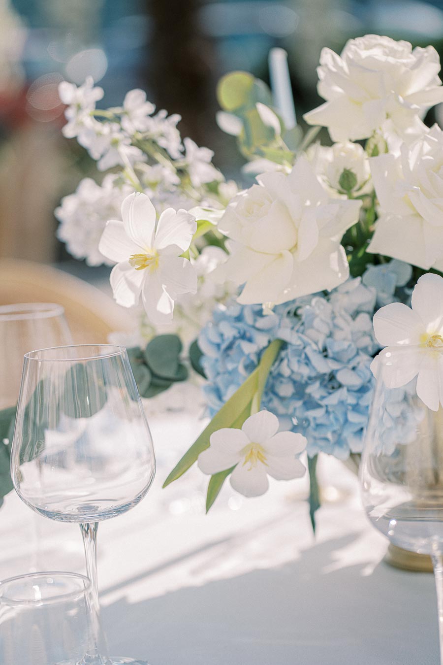 Elegant floral arrangement featuring white roses, delicate white flowers, and soft blue hydrangeas on a beautifully set table with clear wine glasses, exuding a serene and romantic atmosphere.