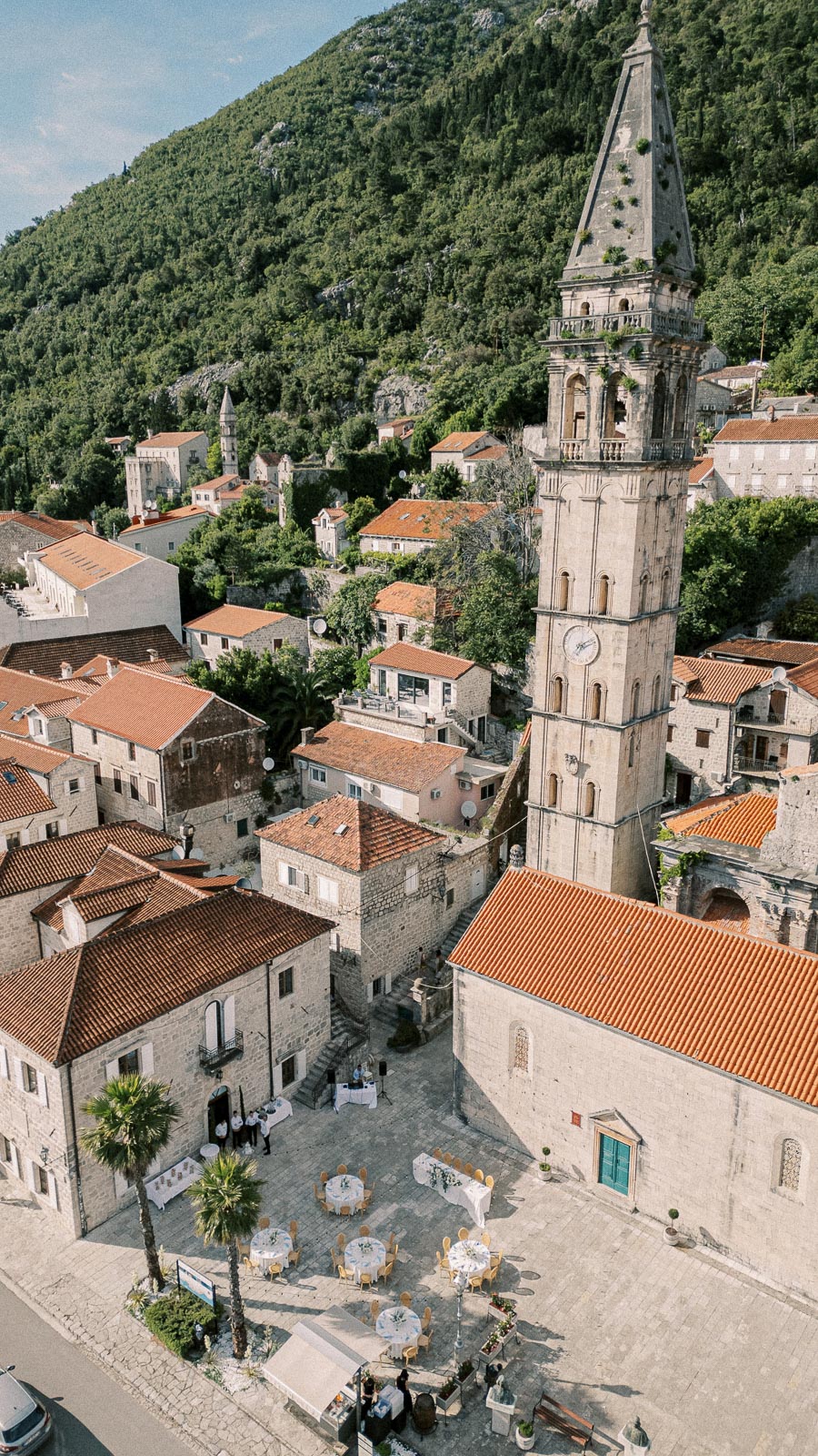 Aerial view of a picturesque Mediterranean town featuring a historic stone bell tower with a clock, surrounded by charming buildings with terracotta roofs. The town is set against a lush green hillside, with a small courtyard below set for an outdoor event, including round tables with white tablecloths and chairs.