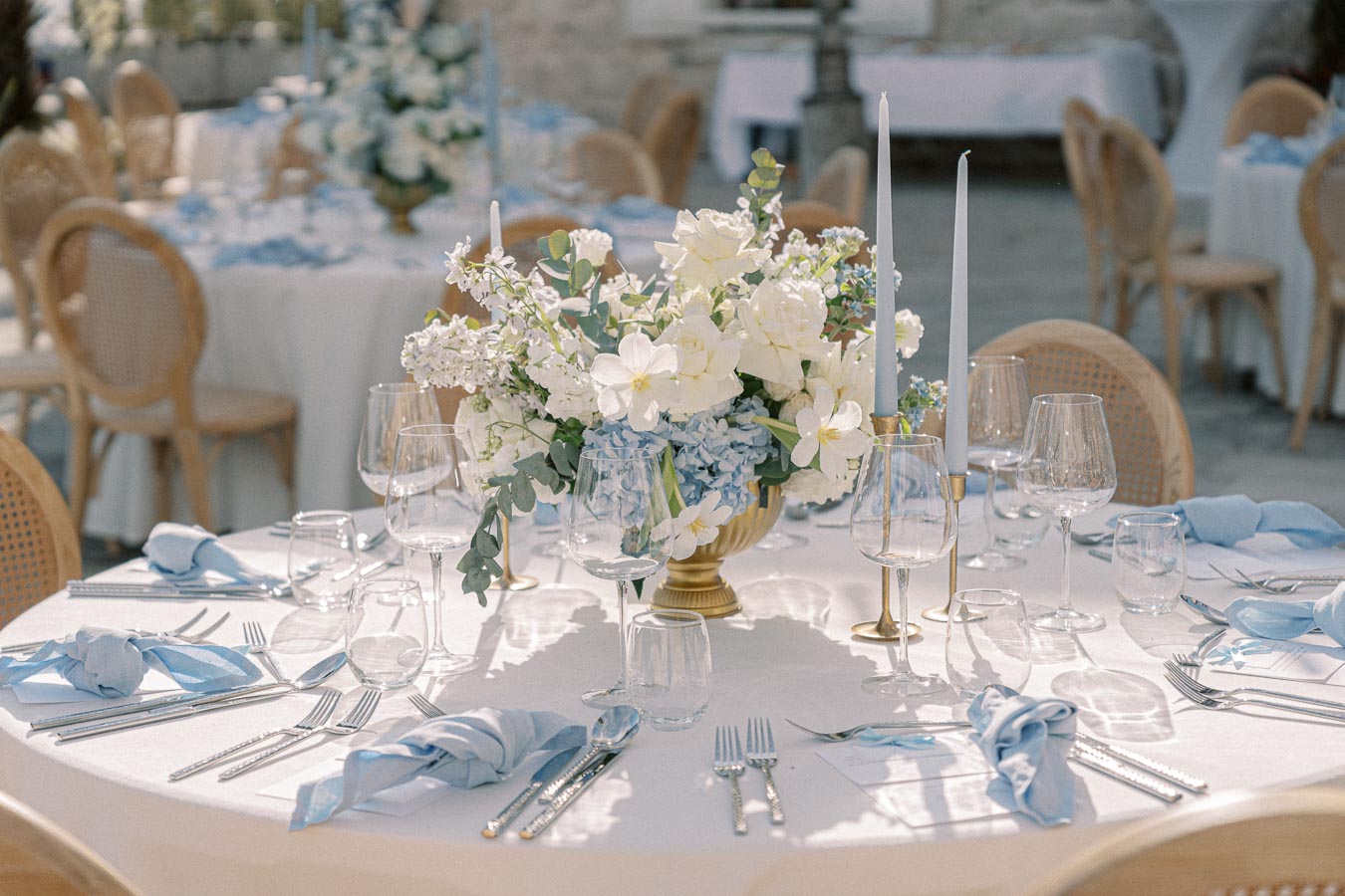 Elegant wedding reception table set up with a white tablecloth, adorned with a centerpiece of white and blue flowers, tall candlesticks, crystal glasses, and neatly arranged silverware, creating a sophisticated and romantic outdoor dining ambiance.