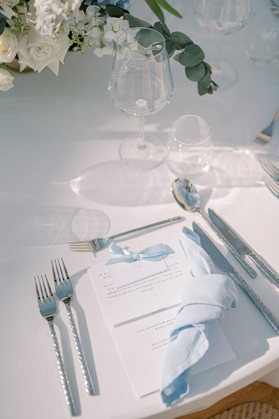 Elegant wedding table setting featuring a white tablecloth, polished silverware, a neatly folded light blue napkin, a menu card with a blue ribbon, and delicate white and green floral centerpiece.