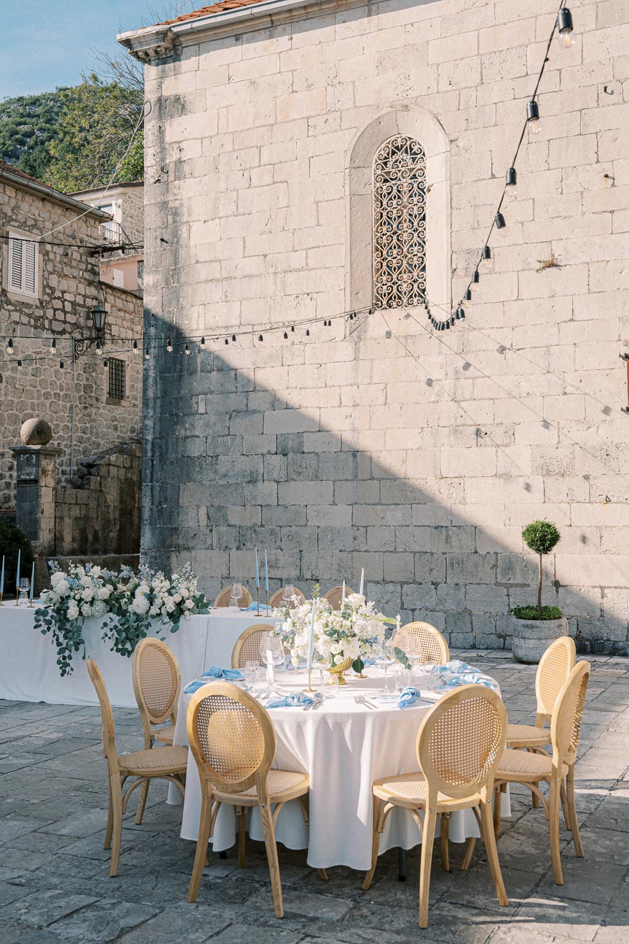 Elegant outdoor dining setup in a rustic courtyard, featuring a round table with wooden chairs, white tablecloth, and floral centerpieces, surrounded by stone walls and decorative string lights.