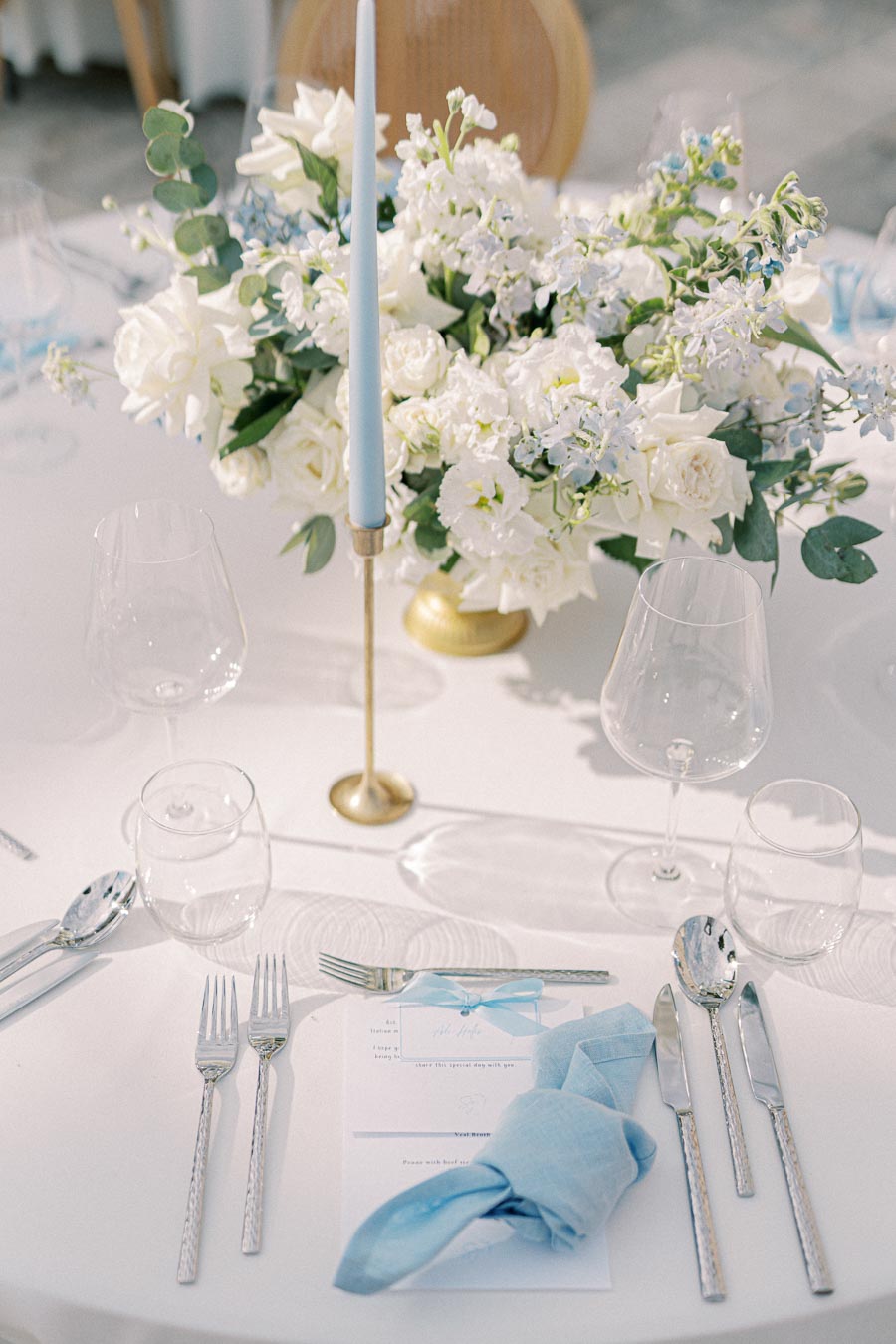 Elegant wedding table setting with white floral centerpiece, light blue taper candle, and tableware, including a blue napkin and glassware, on a white tablecloth.