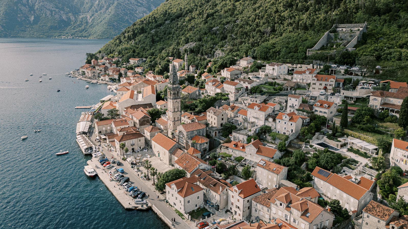 Aerial view of a picturesque coastal village with traditional stone houses and red-tiled roofs nestled by the Adriatic Sea, surrounded by lush green hills. Row boats and yachts are docked along the waterfront, highlighting the charming Mediterranean architecture and serene natural landscape.