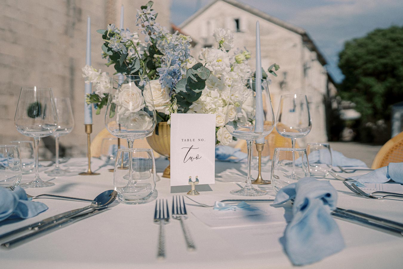 Elegant wedding table setting with white floral centerpiece, blue napkins, and glassware, labeled as Table Number Two in an outdoor setting.