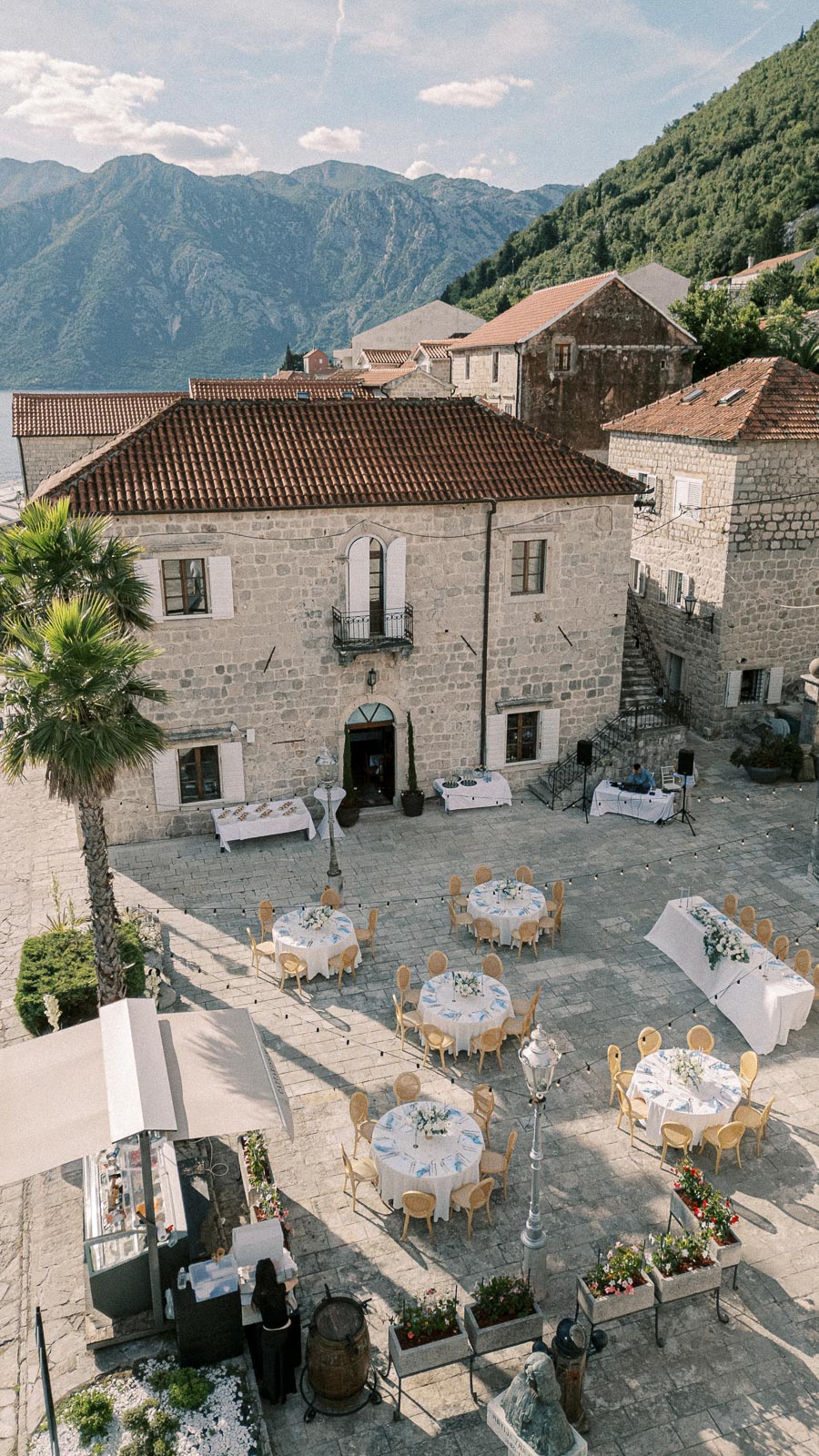 Historic stone courtyard set up for an outdoor event, featuring round tables with white tablecloths and floral centerpieces, surrounded by mountains and lush greenery.