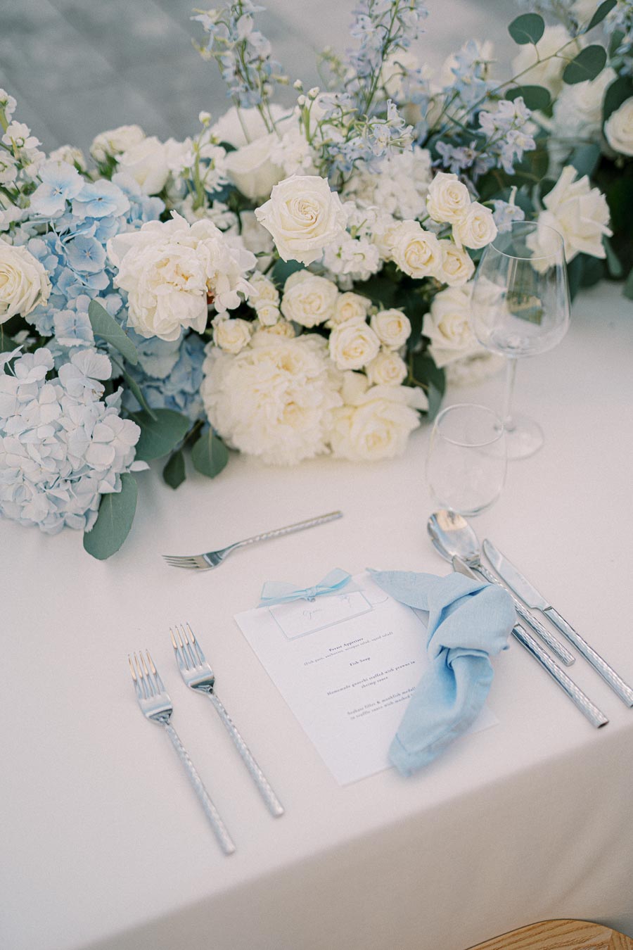 Elegant wedding table setting featuring a white and blue floral centerpiece with roses and hydrangeas, accompanied by a neatly folded blue napkin, menu, and polished silverware on a white tablecloth.