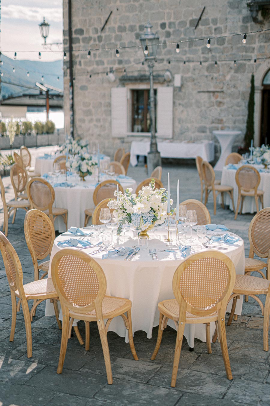Elegant outdoor wedding reception setup with round tables adorned with white tablecloths, floral centerpieces, and blue napkins, surrounded by wooden chairs, against a rustic stone building backdrop.