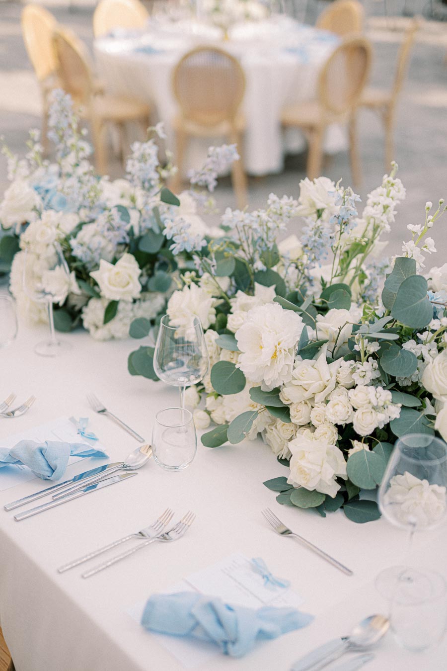 Elegant wedding reception table with white and green floral centerpiece, blue napkins, glassware, and cutlery set, with blurred seating area in the background.