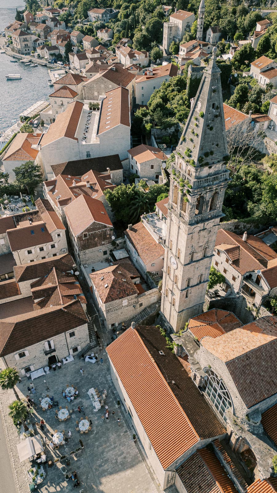 Aerial view of a picturesque coastal town with historic stone buildings and red-tiled roofs, featuring a prominent bell tower surrounded by greenery. Scenic waterfront with boats docked along the shoreline.