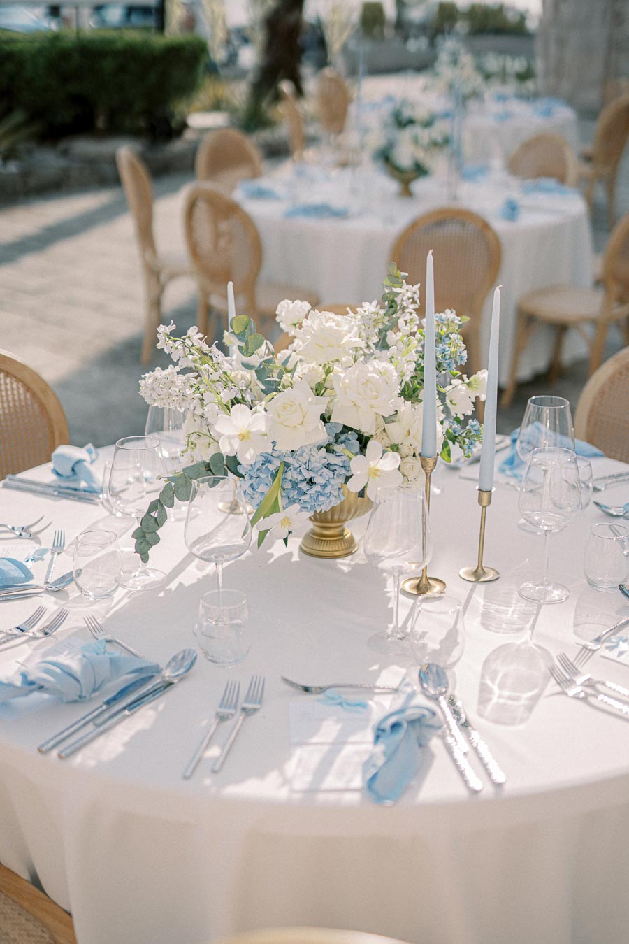 Elegant outdoor wedding table setting with white and blue floral centerpiece, gold candlesticks, and delicate glassware under natural sunlight.