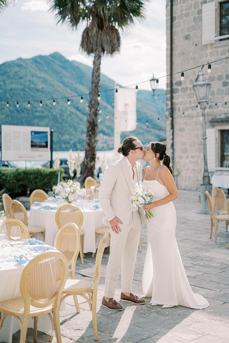 Bride and groom sharing a kiss at an elegant outdoor wedding venue by the sea, surrounded by floral arrangements and scenic mountain views.