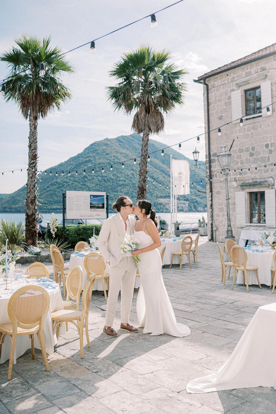 Bride and groom sharing a kiss at an outdoor wedding venue, set against a backdrop of palm trees and scenic mountains, with elegant table settings arranged for a reception.