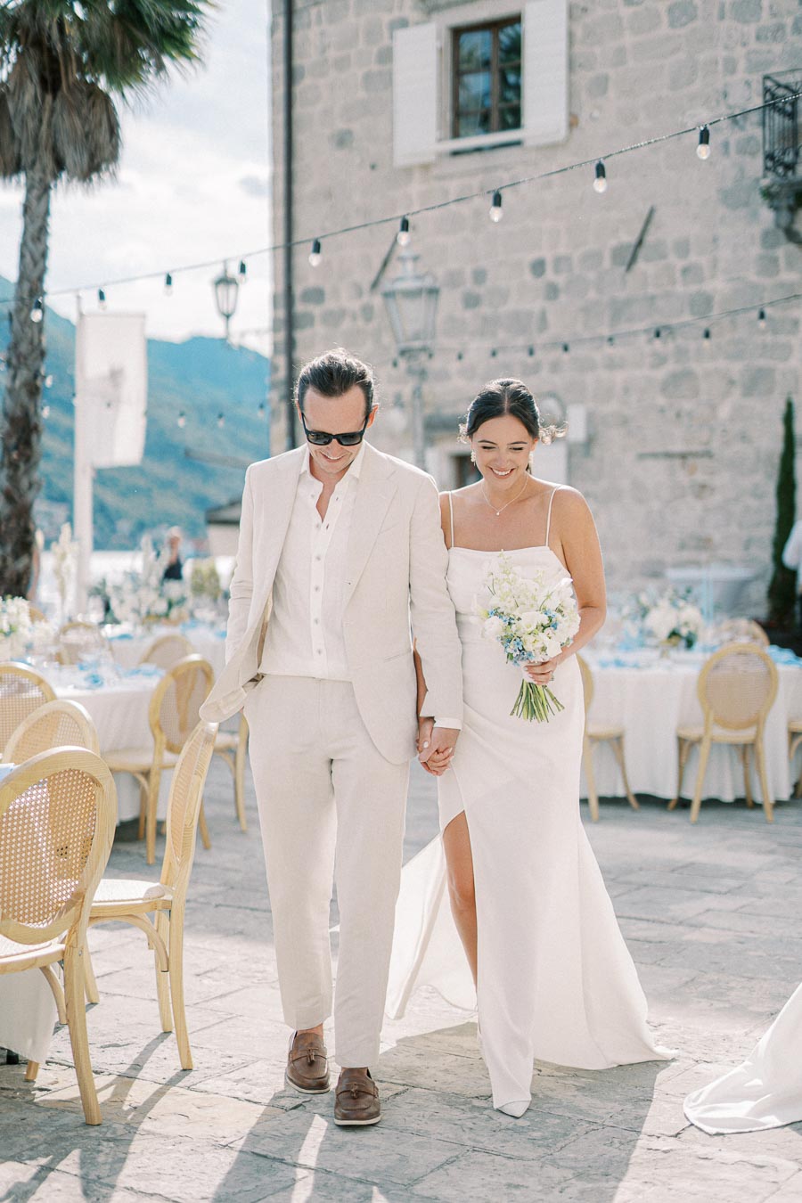 Elegant couple walking hand in hand at an outdoor wedding, featuring a bride in a stylish white gown holding a bouquet, and a groom in a light suit with sunglasses, surrounded by charming patio decor and scenic mountainous backdrop.