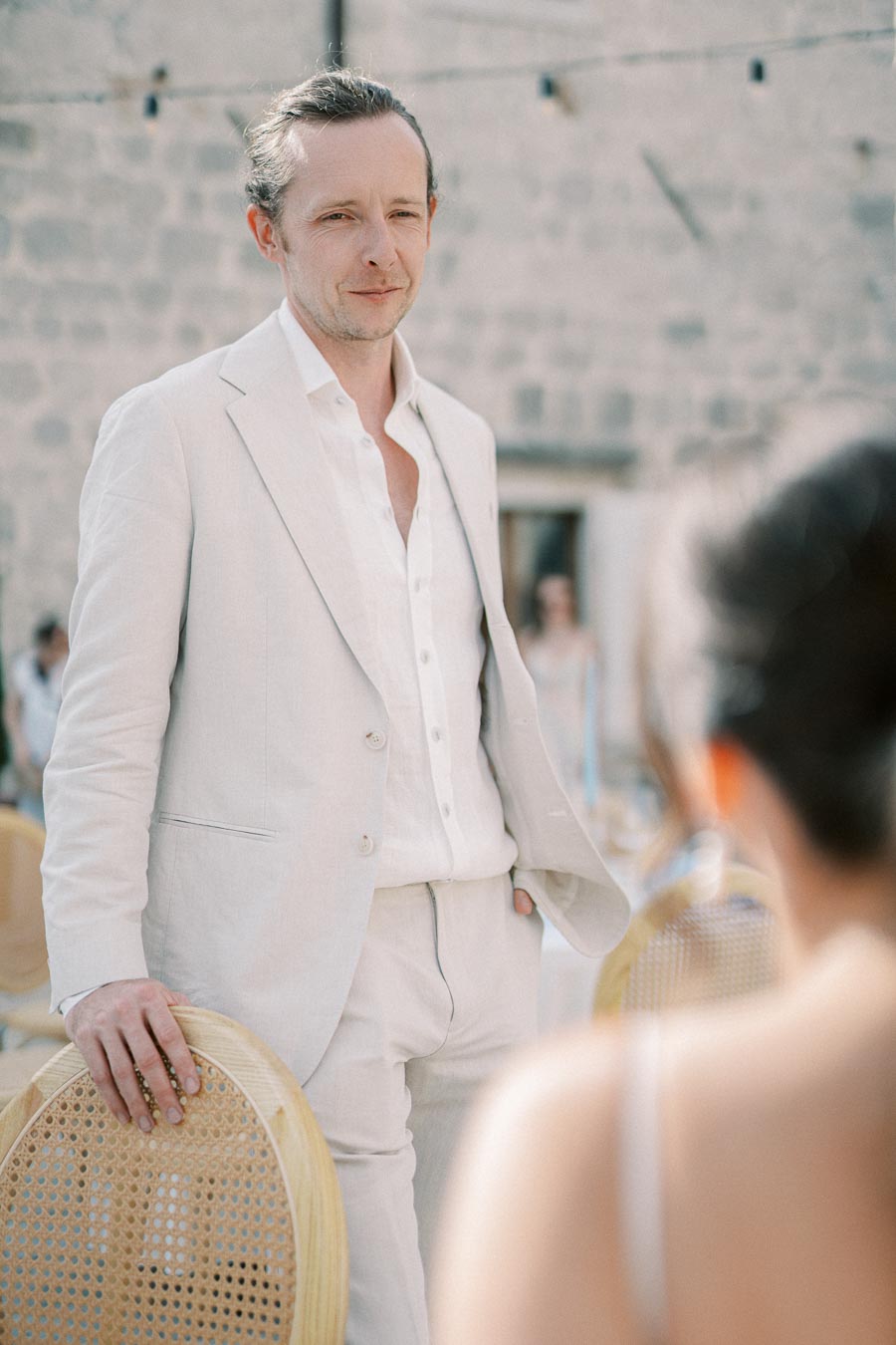 A man in a light suit standing outdoors near a stone wall, casually resting his hand on a wicker chair, with people in the background out of focus.
