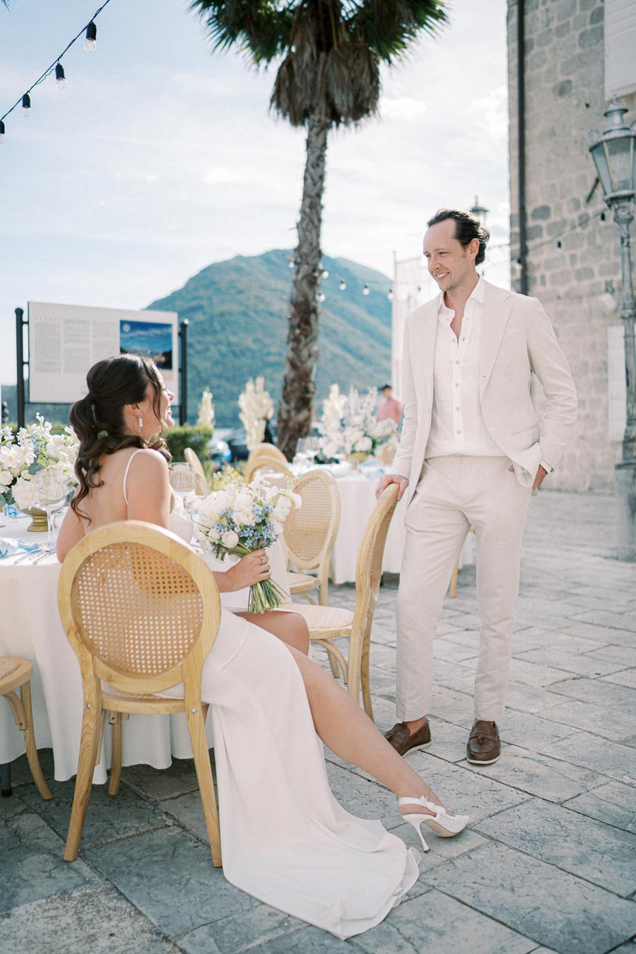 Elegant outdoor wedding scene with a bride in a white dress holding a bouquet, seated at a decorated table, and a groom in a light suit standing and smiling, set against a scenic mountainous backdrop.