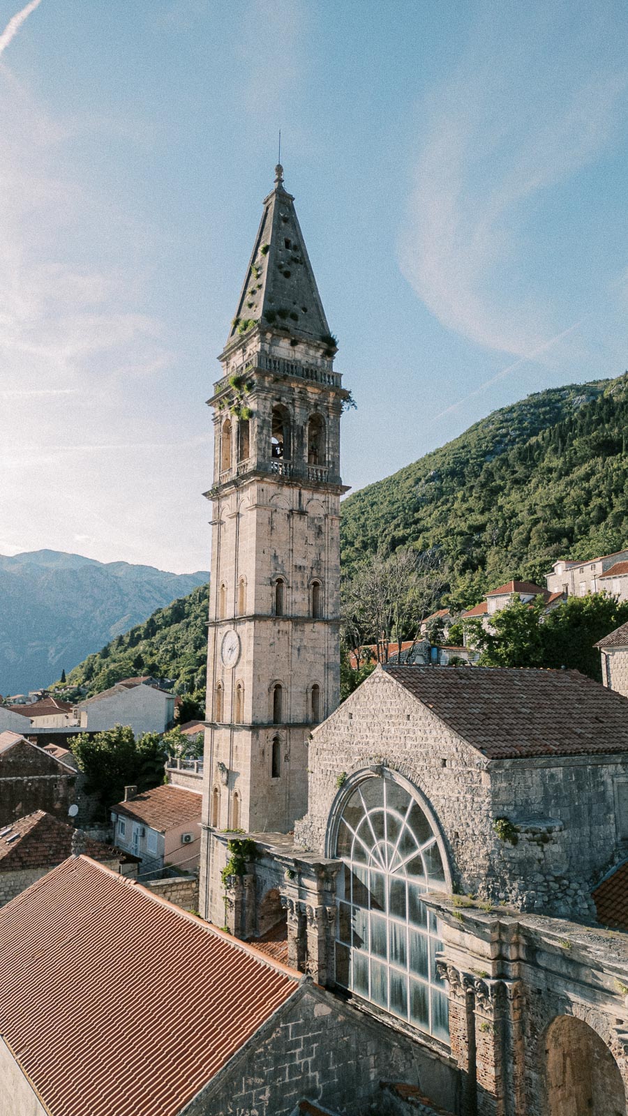 Tall stone clock tower next to a rustic church with arched windows, set against a backdrop of lush green mountains and a clear blue sky.