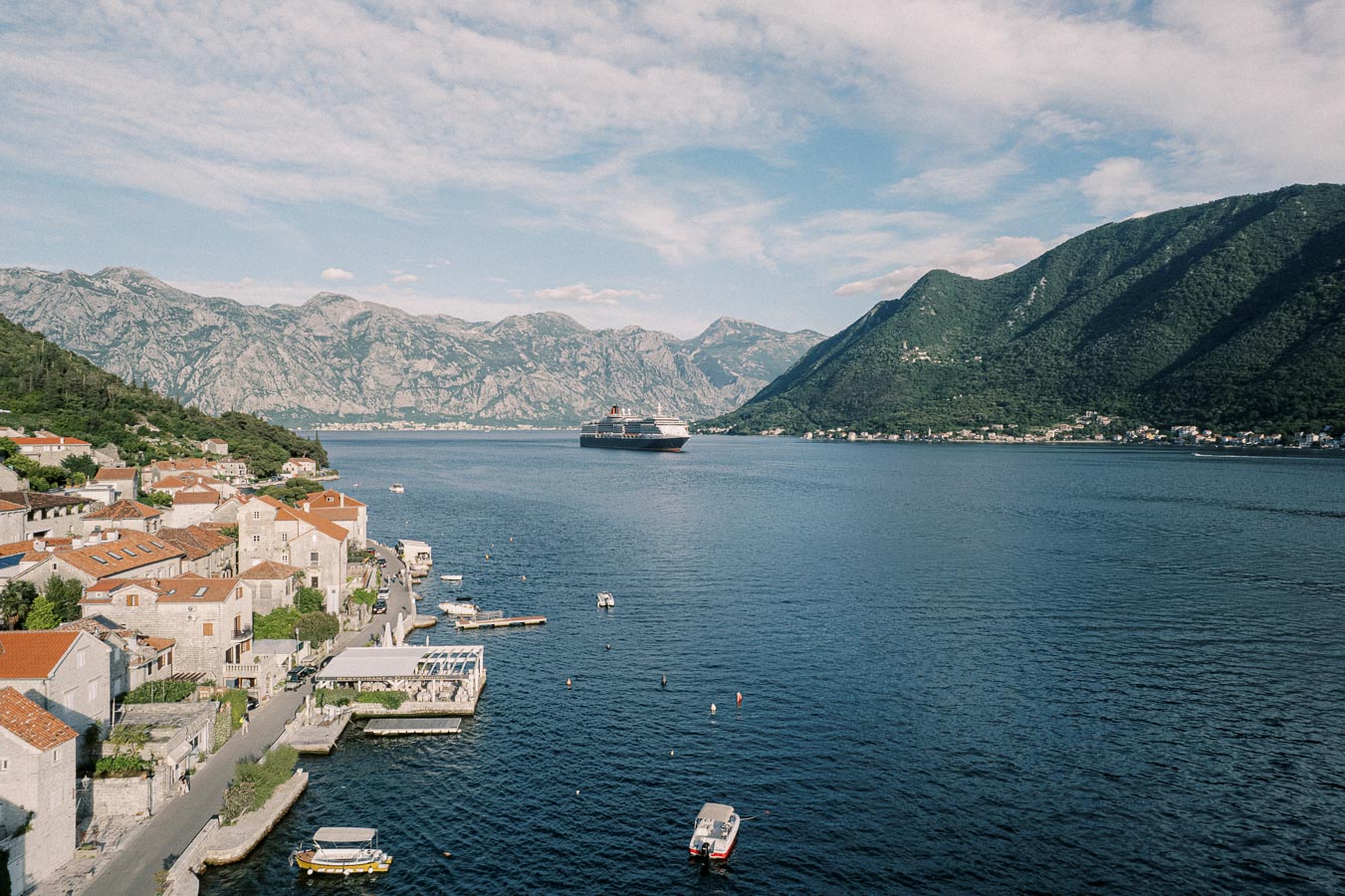 A scenic view of a coastal town with red-roofed buildings along the Adriatic Sea, surrounded by mountainous landscapes, and a cruise ship sailing in the blue waters under a partly cloudy sky.