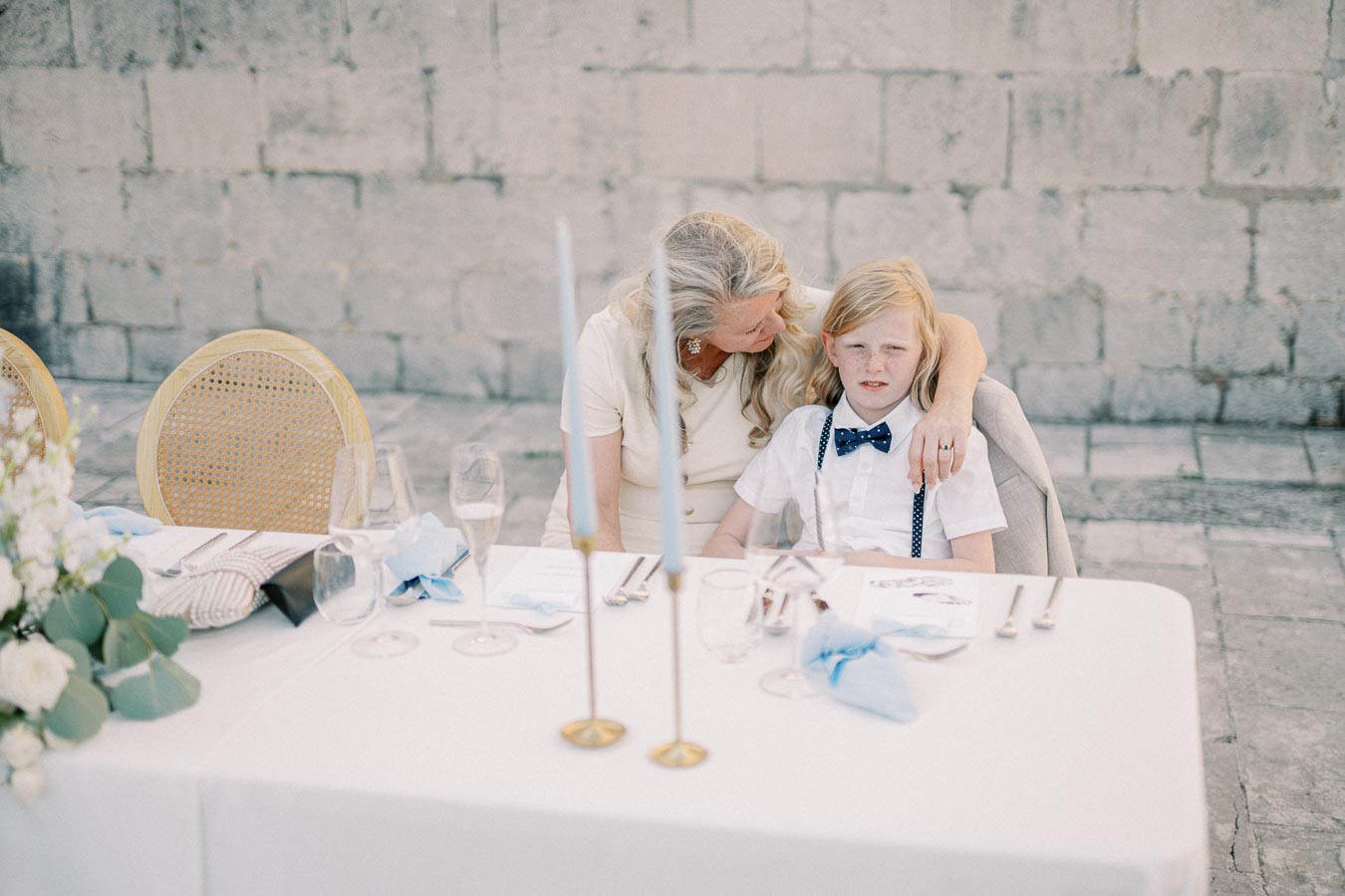 A caring woman comforting a young boy dressed in a white shirt and blue bow tie at an elegant outdoor table setting with candles, glassware, and floral decorations.