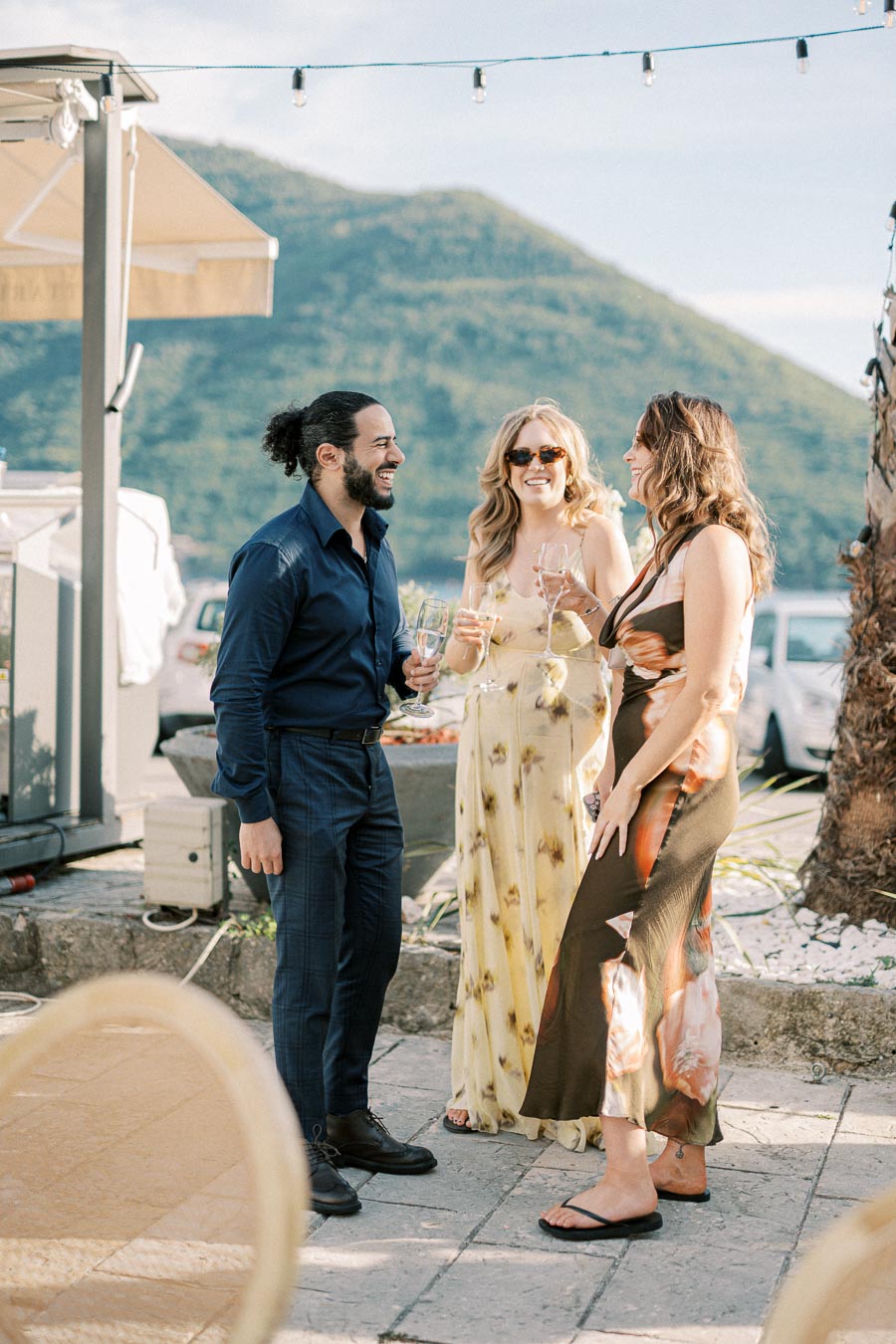 Three people enjoy a social gathering outdoors, dressed in summer attire, holding wine glasses and smiling, with mountains in the background.