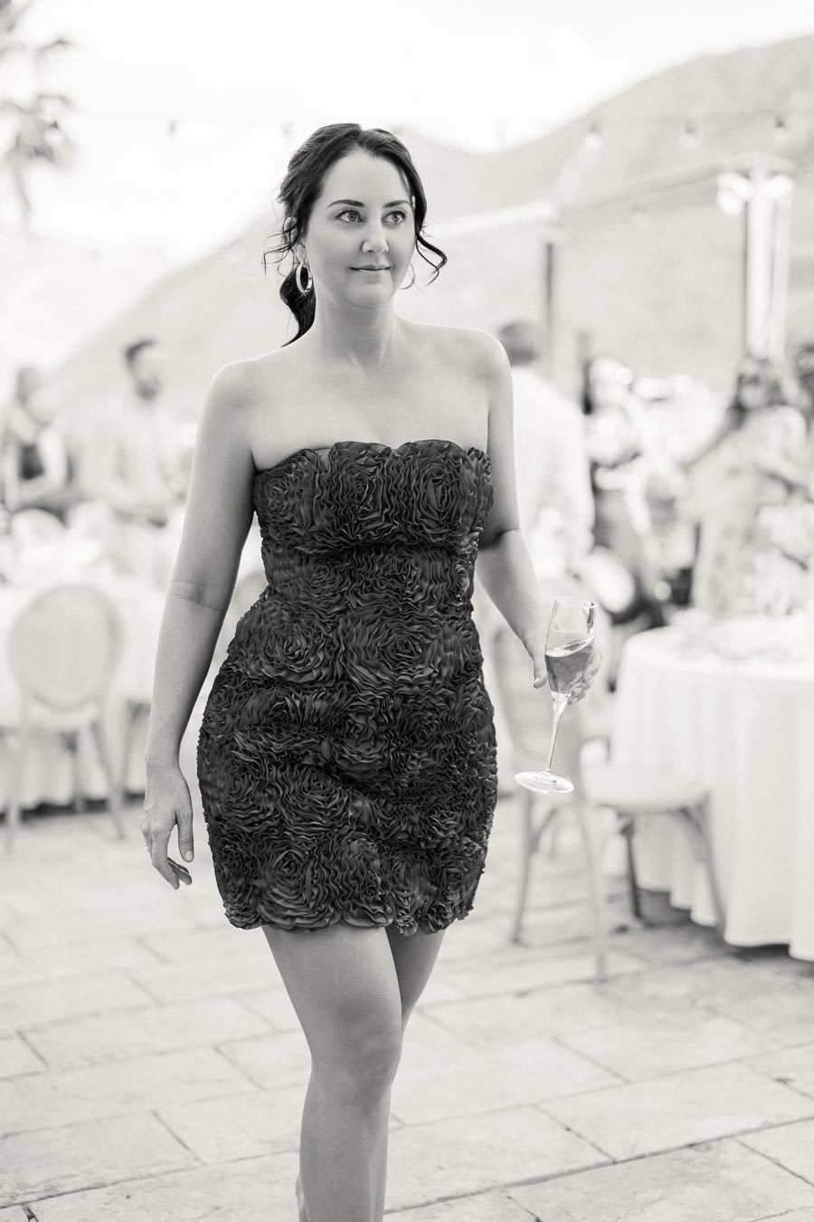 A woman in a strapless, floral-textured dress walks confidently at an outdoor event, holding a champagne glass. The background features seated guests and tables, suggesting a formal or celebratory occasion. Black and white photography captures the elegant atmosphere.