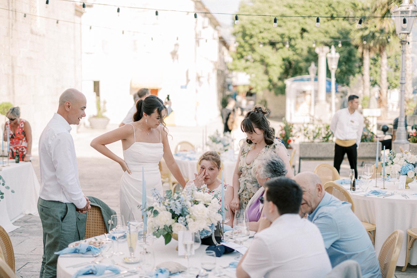 Outdoor wedding reception scene with guests interacting around a decorated table, featuring elegant floral arrangements and pastel-colored linens in a sunlit courtyard.