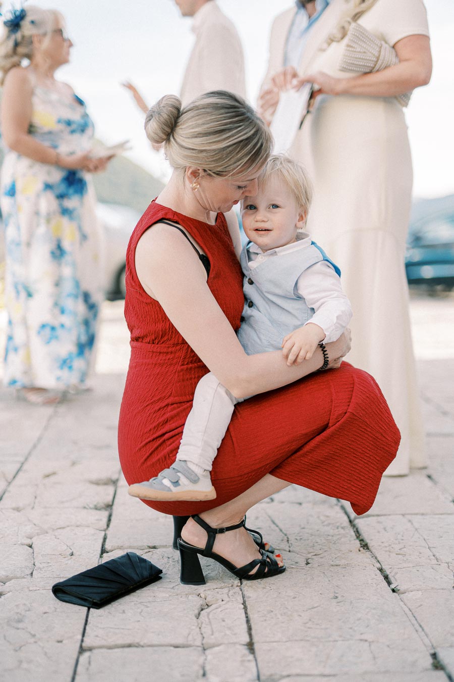 A woman in a red dress lovingly hugs a toddler in a light blue vest while kneeling on a stone path at an outdoor event. Other guests are visible in the background.