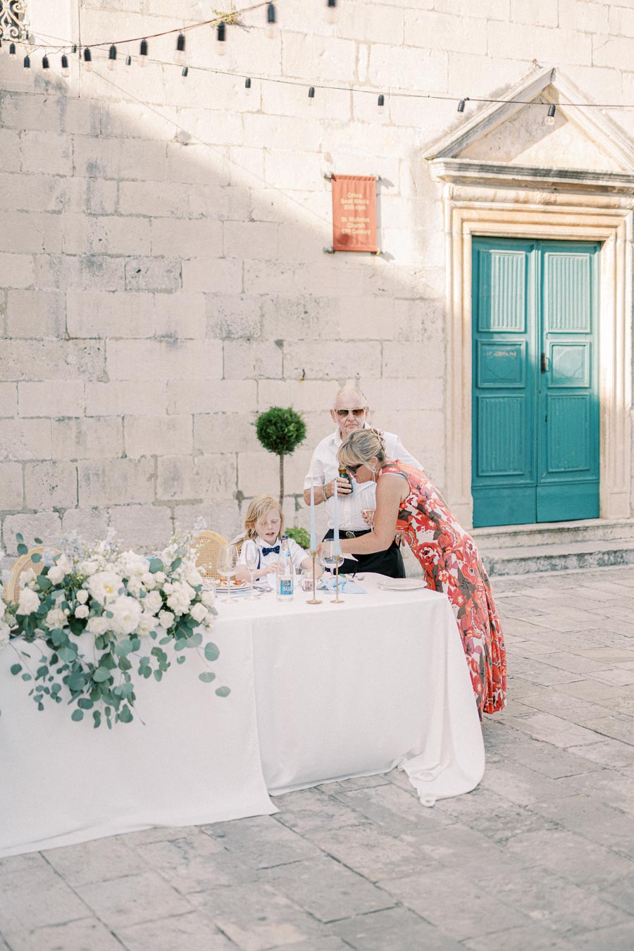 A young child in a formal outfit sits at a beautifully decorated outdoor table with white flowers and greenery, accompanied by two adults interacting with each other against a historic stone wall backdrop and a teal door.