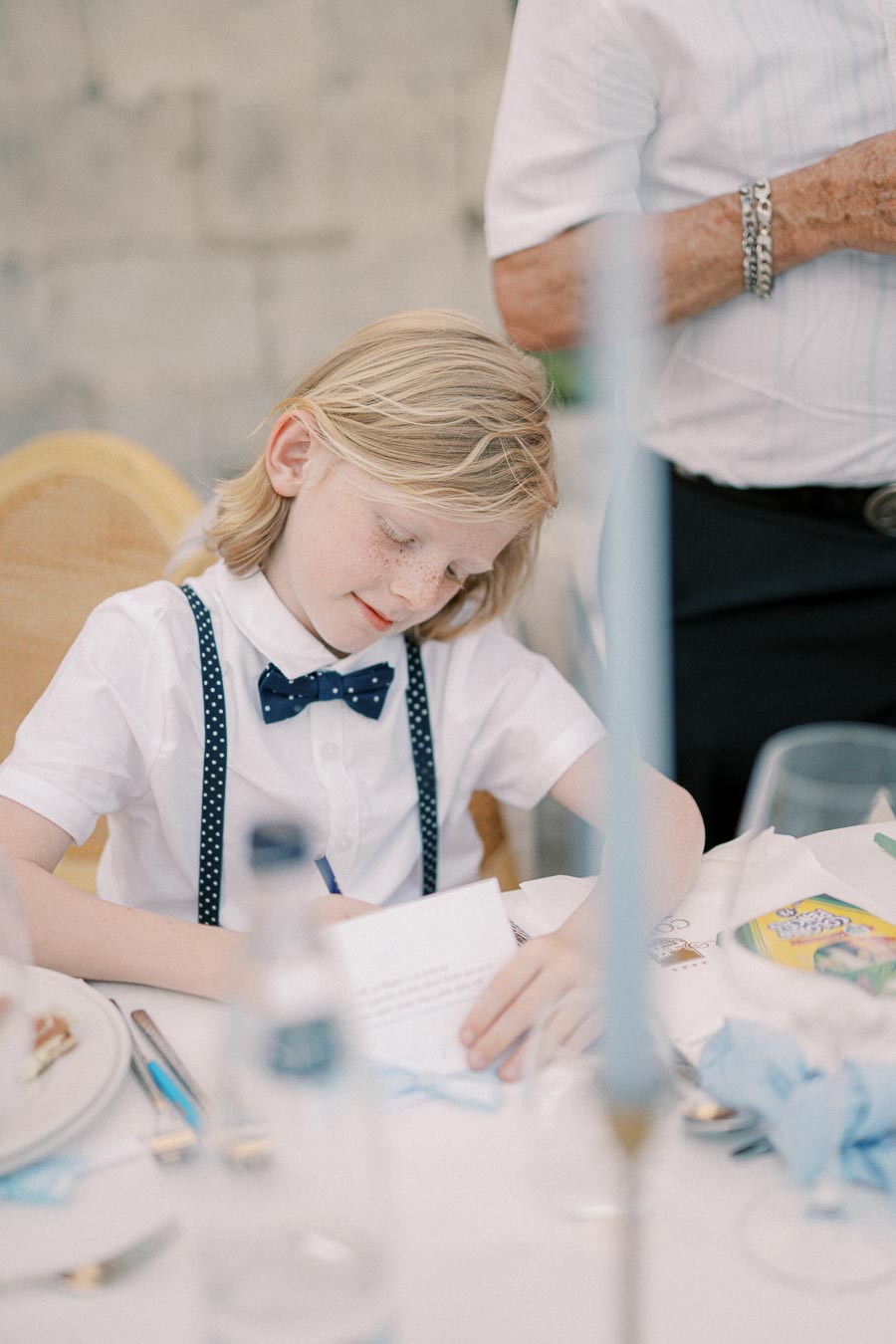 A young boy with blonde hair wearing a white shirt, polka dot suspenders, and a bow tie writes on paper at a table set with dishes and cutlery.