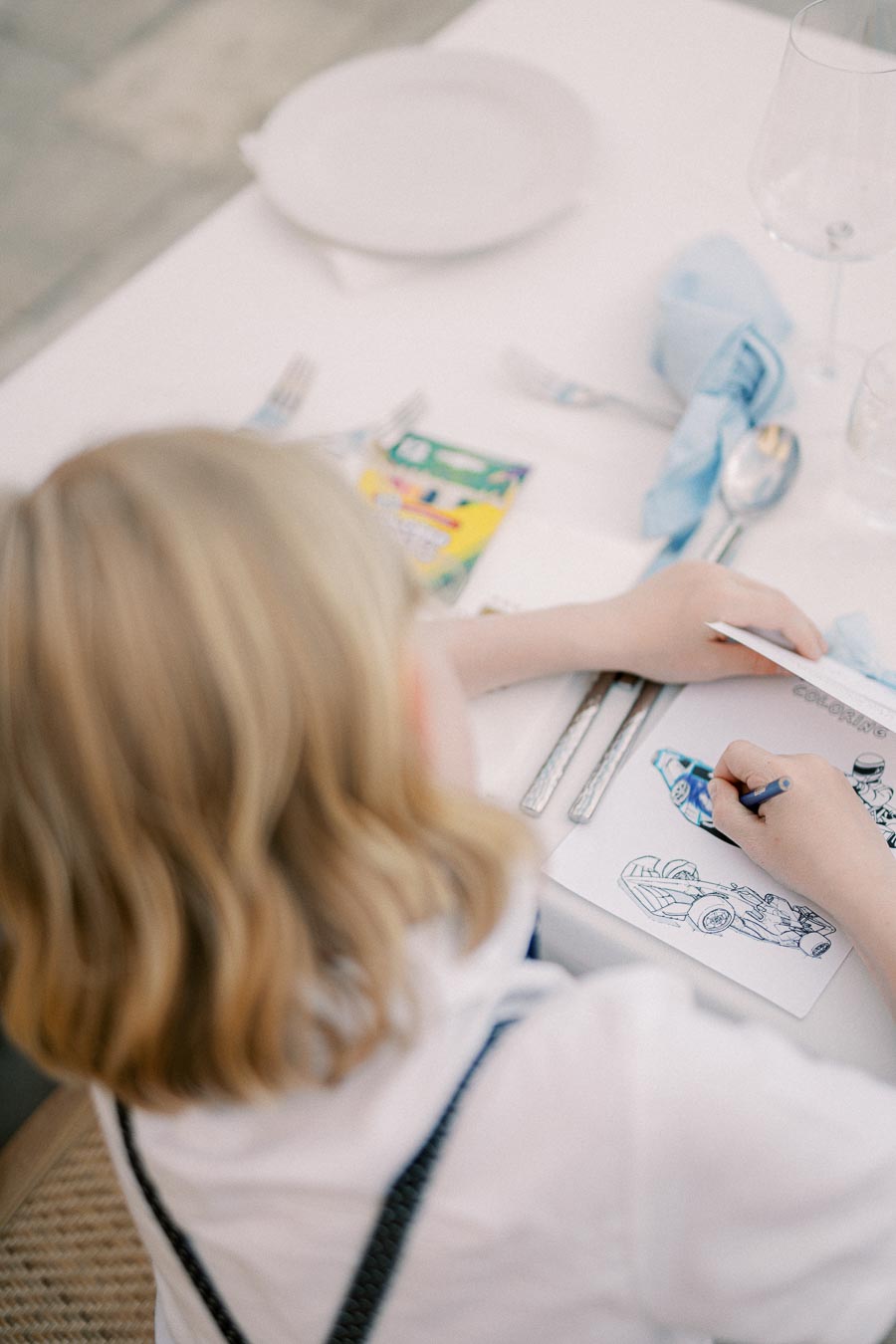 A child coloring a picture of a car at a table set for a meal, with utensils and a glass nearby.
