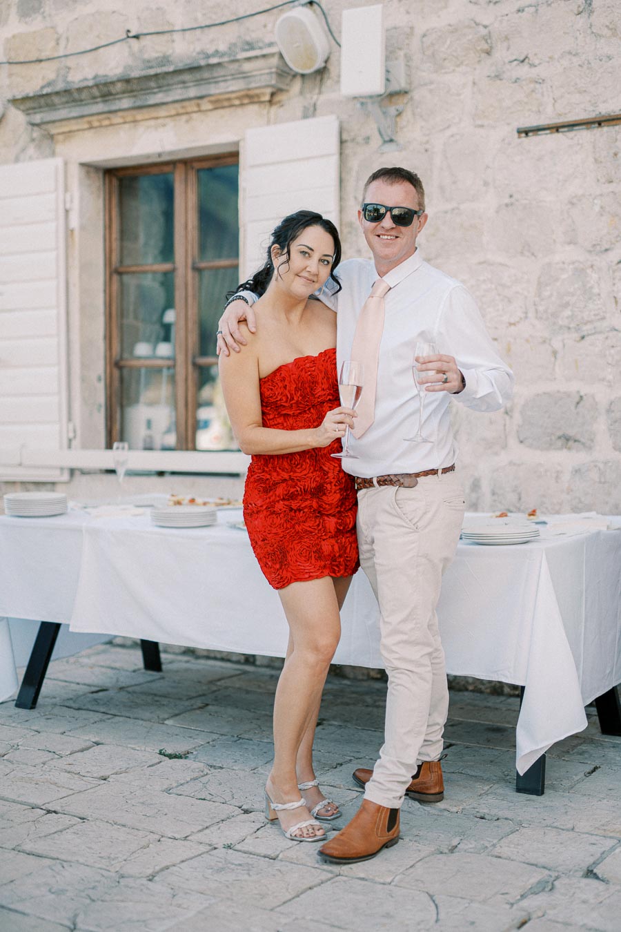 A couple celebrating at an outdoor event, dressed elegantly with glasses of champagne, set against a rustic stone backdrop.