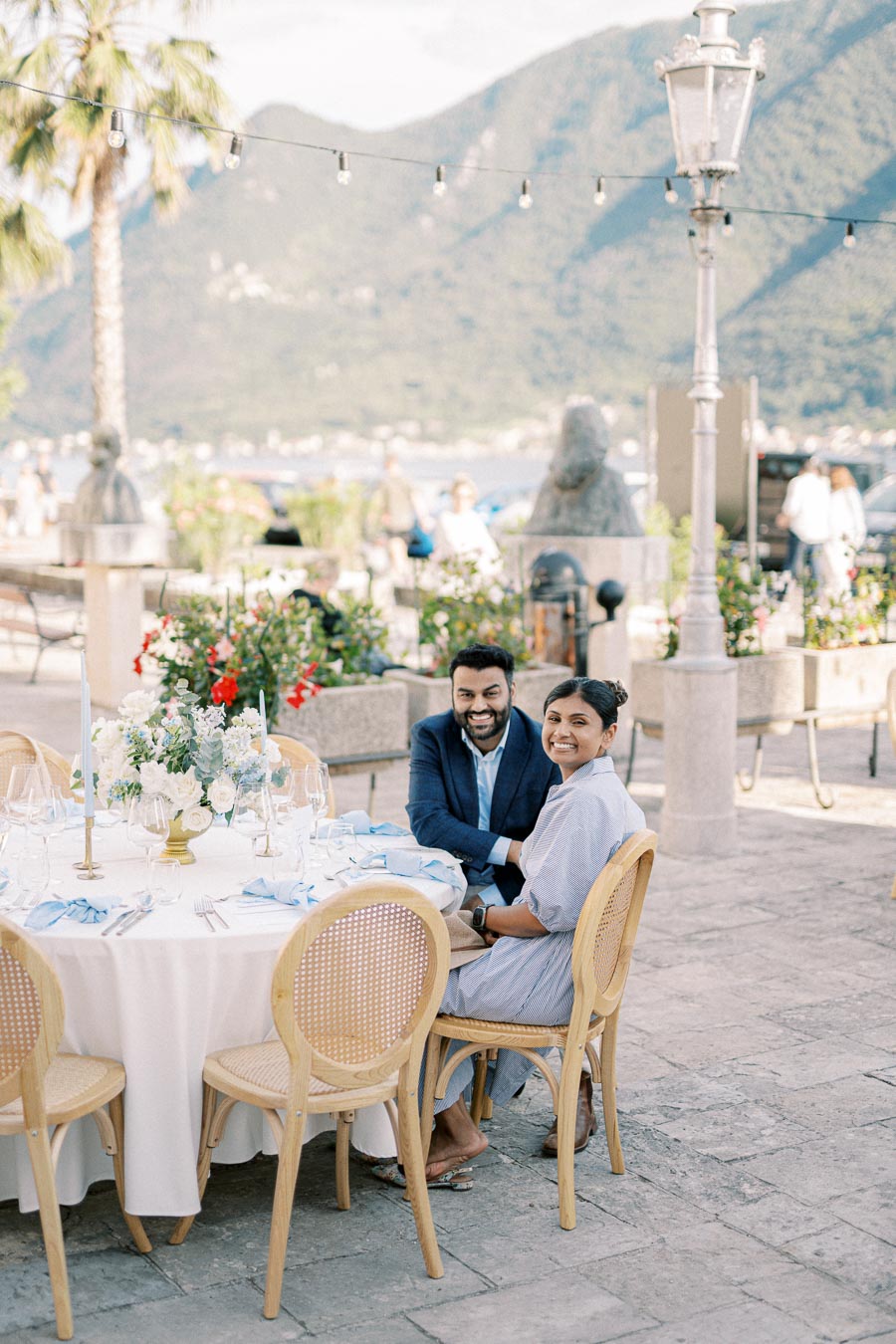 Couple smiling at an elegantly set outdoor dining table with scenic mountain and lake views in the background.