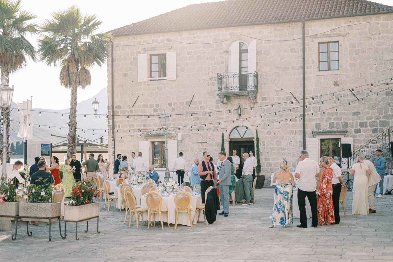 Outdoor wedding reception with guests socializing beside a historic stone building, featuring decorative lighting and elegantly set tables adorned with floral arrangements.