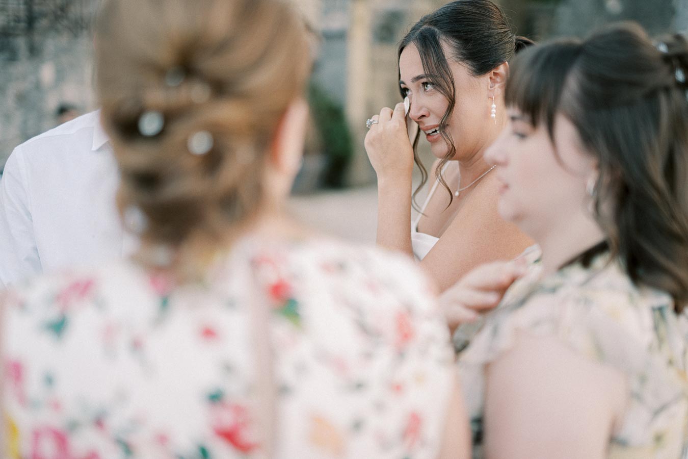 A bride in a white dress wipes a tear of joy surrounded by friends in floral dresses during an emotional wedding moment.