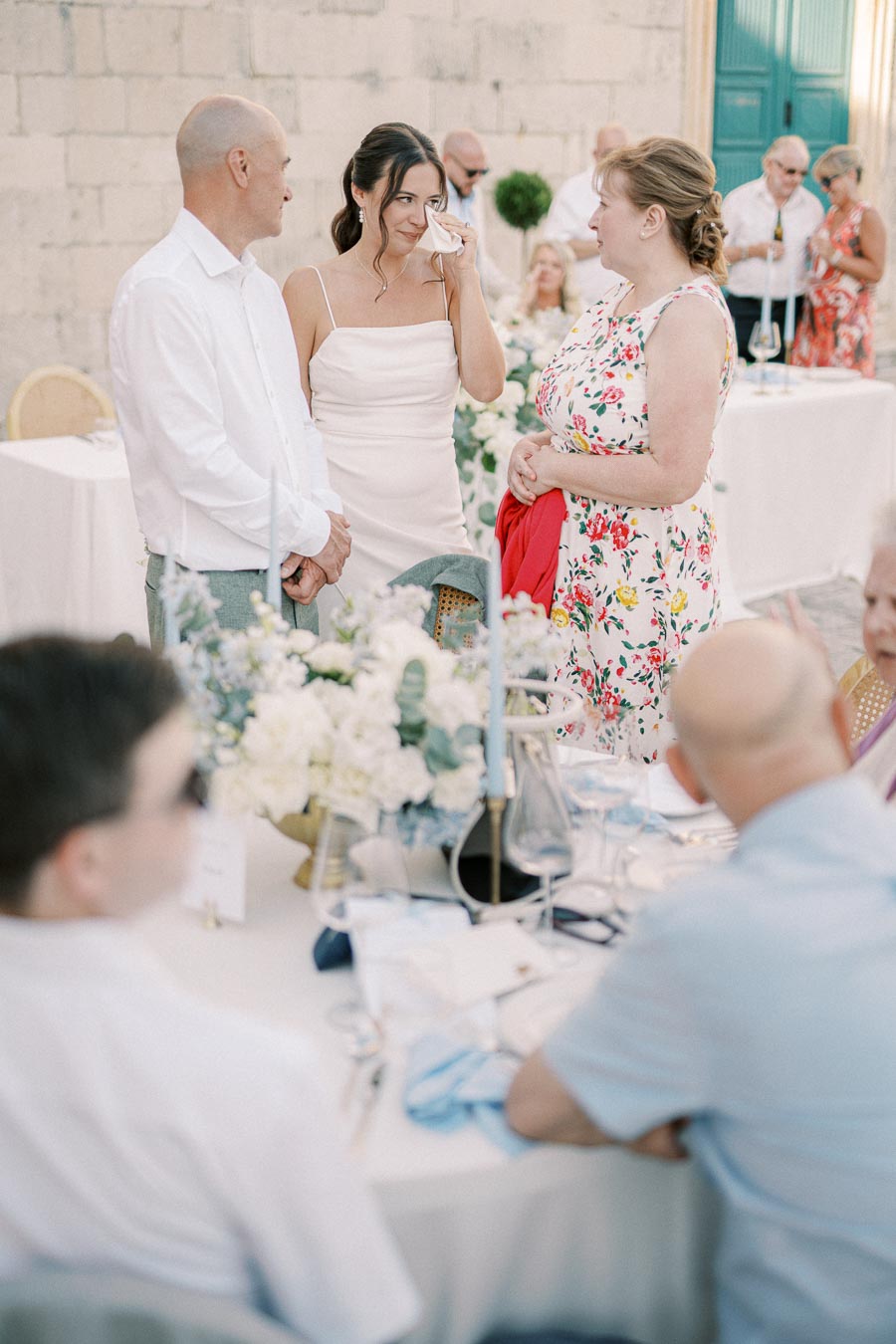 A bride in a white dress wipes her tears with a tissue while standing with guests at an outdoor wedding reception, surrounded by floral decor and blurred guests in the background.