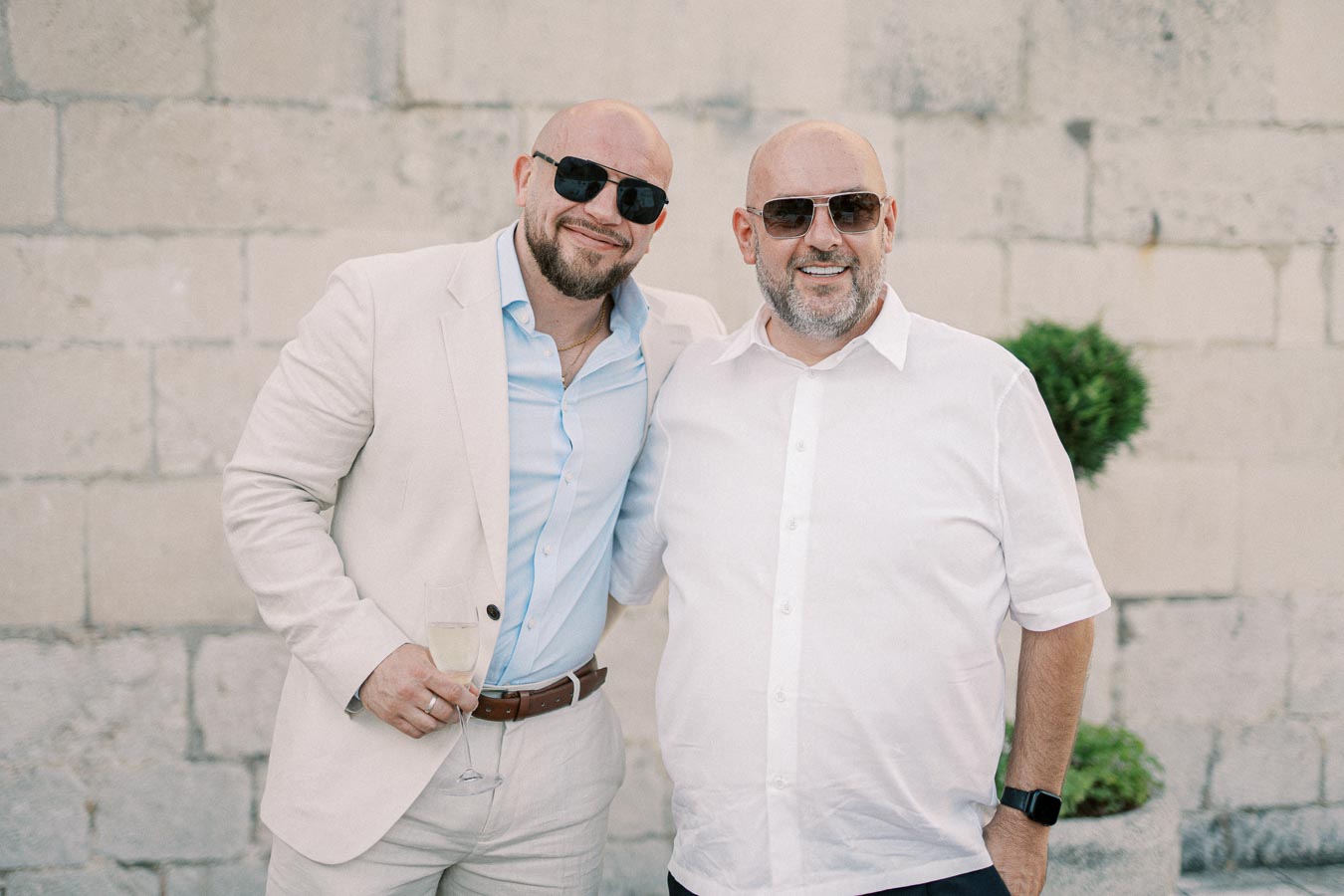 Two smiling men wearing sunglasses and light-colored shirts stand together in front of a textured stone wall, with one holding a glass of champagne and a small plant visible in the background.