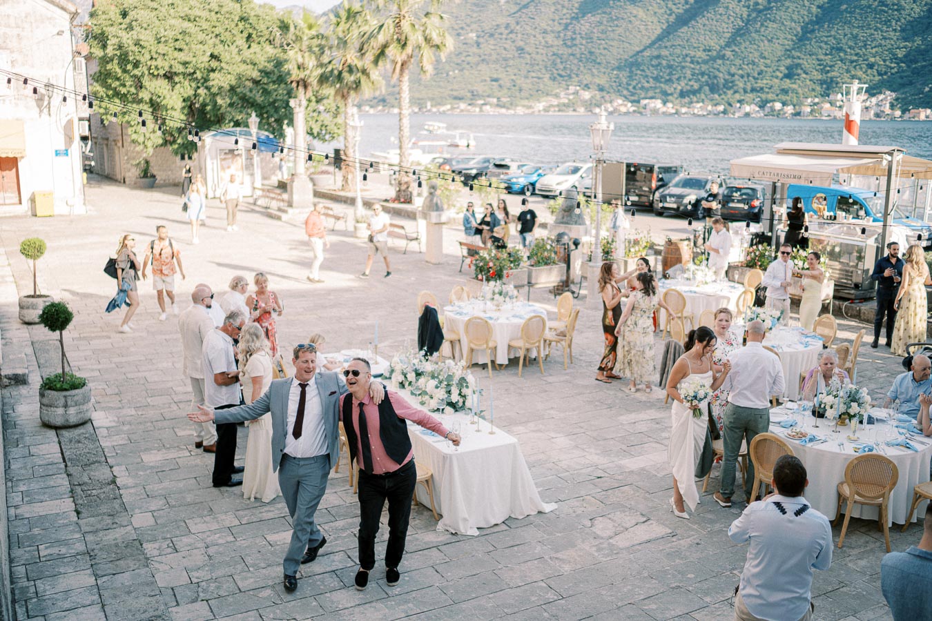 Outdoor wedding reception by a scenic lakeside with guests socializing at elegantly set tables, surrounded by mountains and greenery.