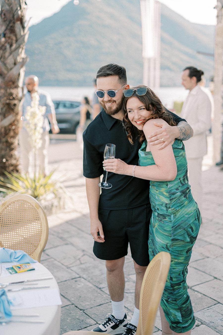 Couple enjoying an outdoor event near a mountain lake, with the woman in a green dress hugging a man in sunglasses holding a champagne glass.