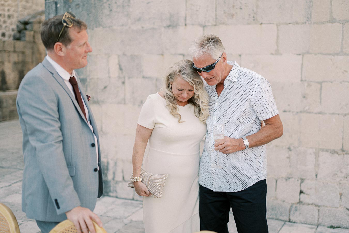 Three people dressed in formal attire at an outdoor event, standing next to a stone wall; two are engaged in conversation while one holds a champagne glass.