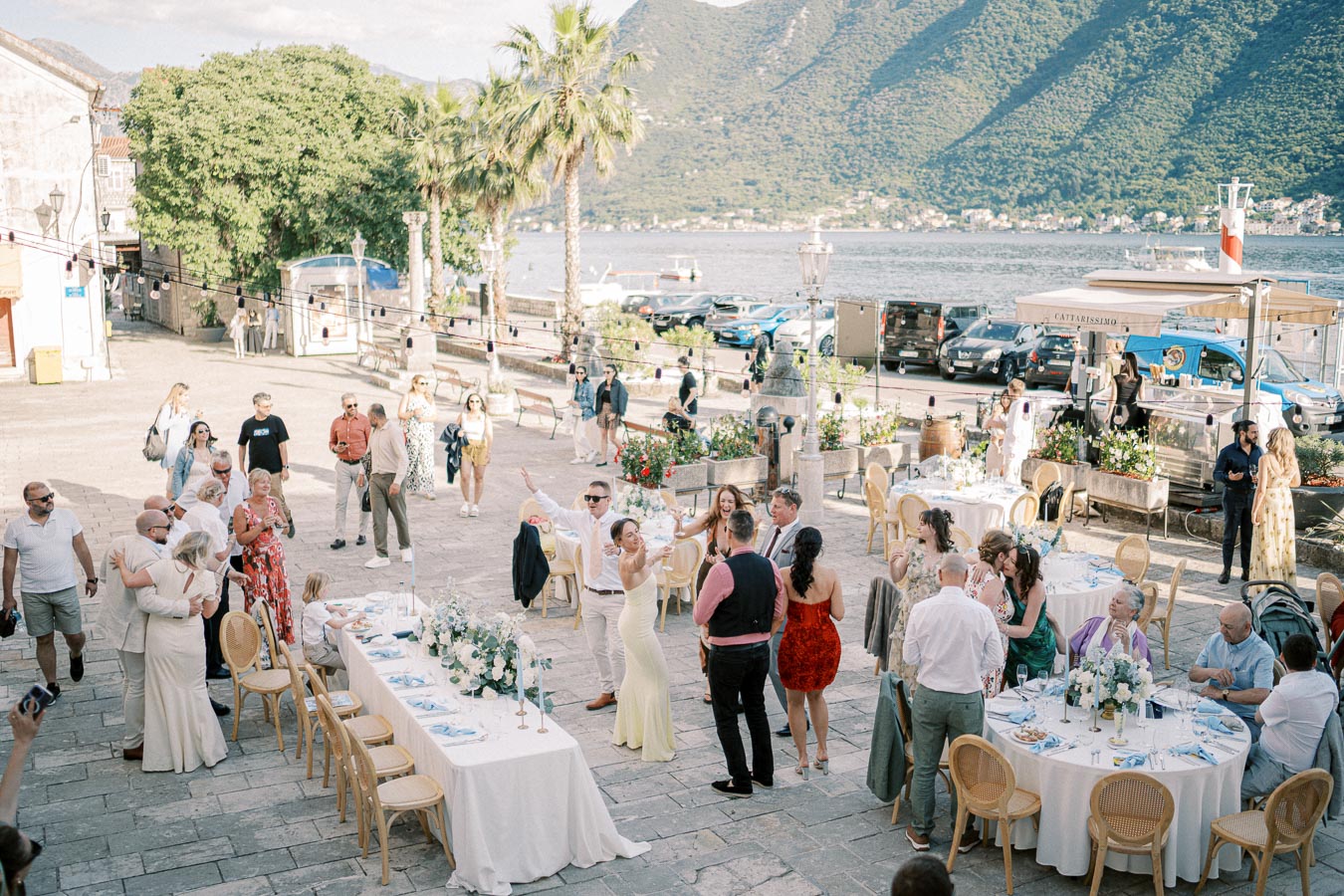 Outdoor wedding reception by the sea, featuring guests mingling around elegantly decorated tables, with mountains and water in the background.