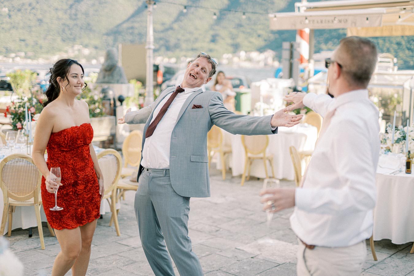 A joyful group of wedding guests dancing and celebrating outdoors, with a mountainous landscape in the background. One person is wearing a red dress, another in a grey suit, and a third in a white shirt, all holding glasses and smiling. Tables with decorations are visible, indicating a festive event.