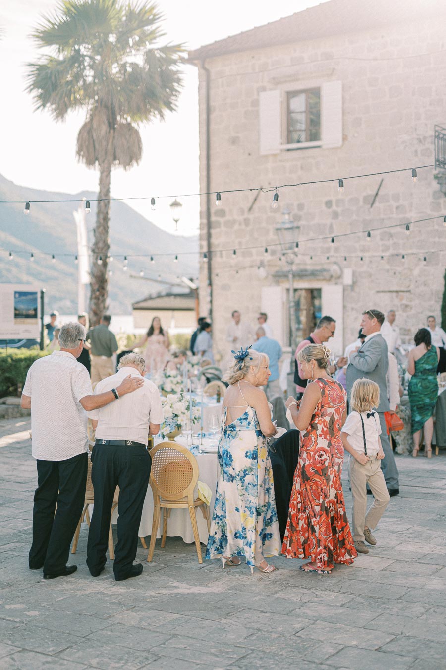 Outdoor wedding reception with guests mingling under string lights near a stone building, palm tree, and mountains in the background.