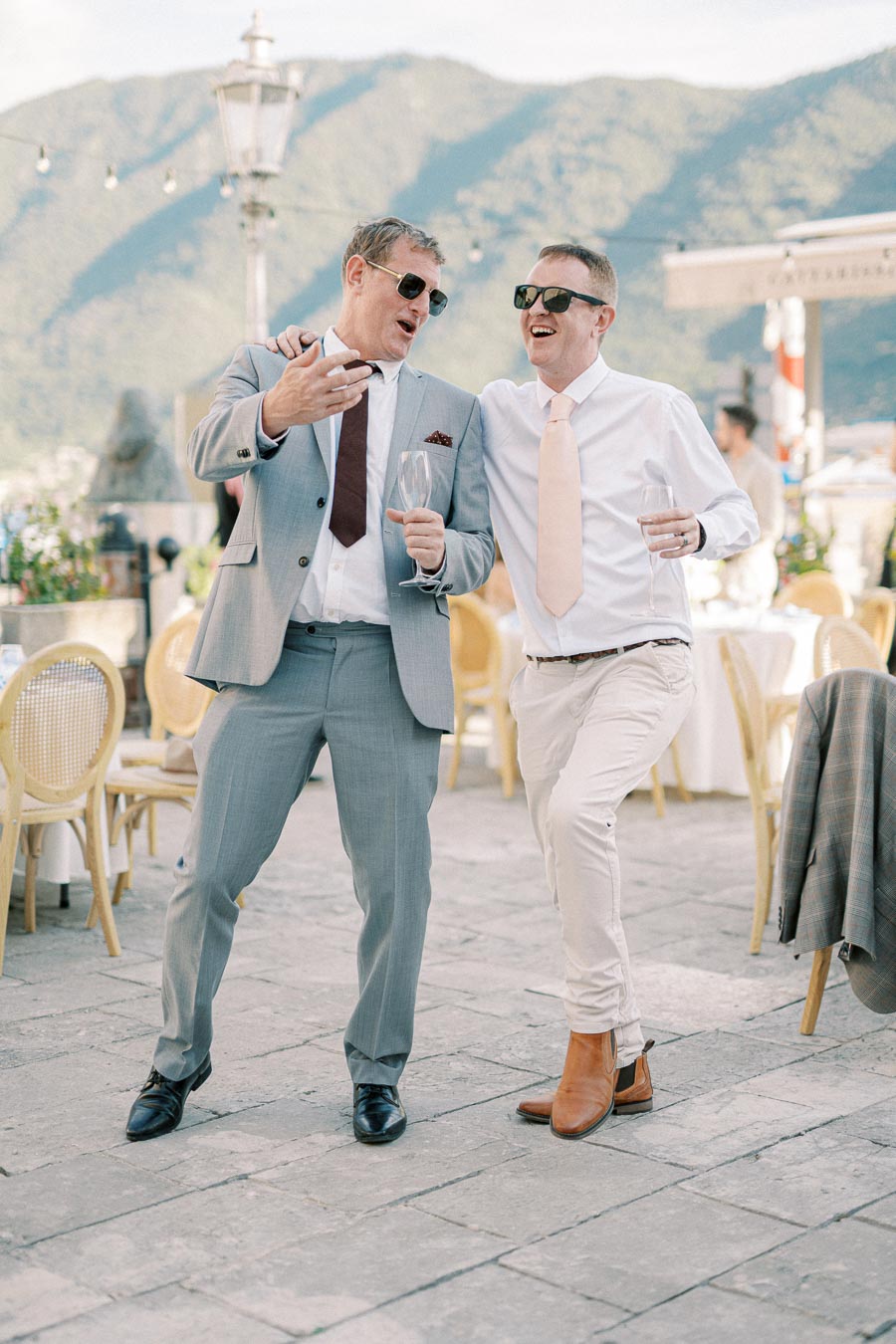 Two men in suits laughing and enjoying a celebration outdoors, with mountains in the background. They are holding drinks and wearing sunglasses, creating a cheerful and relaxed atmosphere.